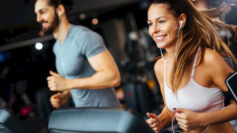 Two people are running on treadmills in a gym, looking happy and focused.