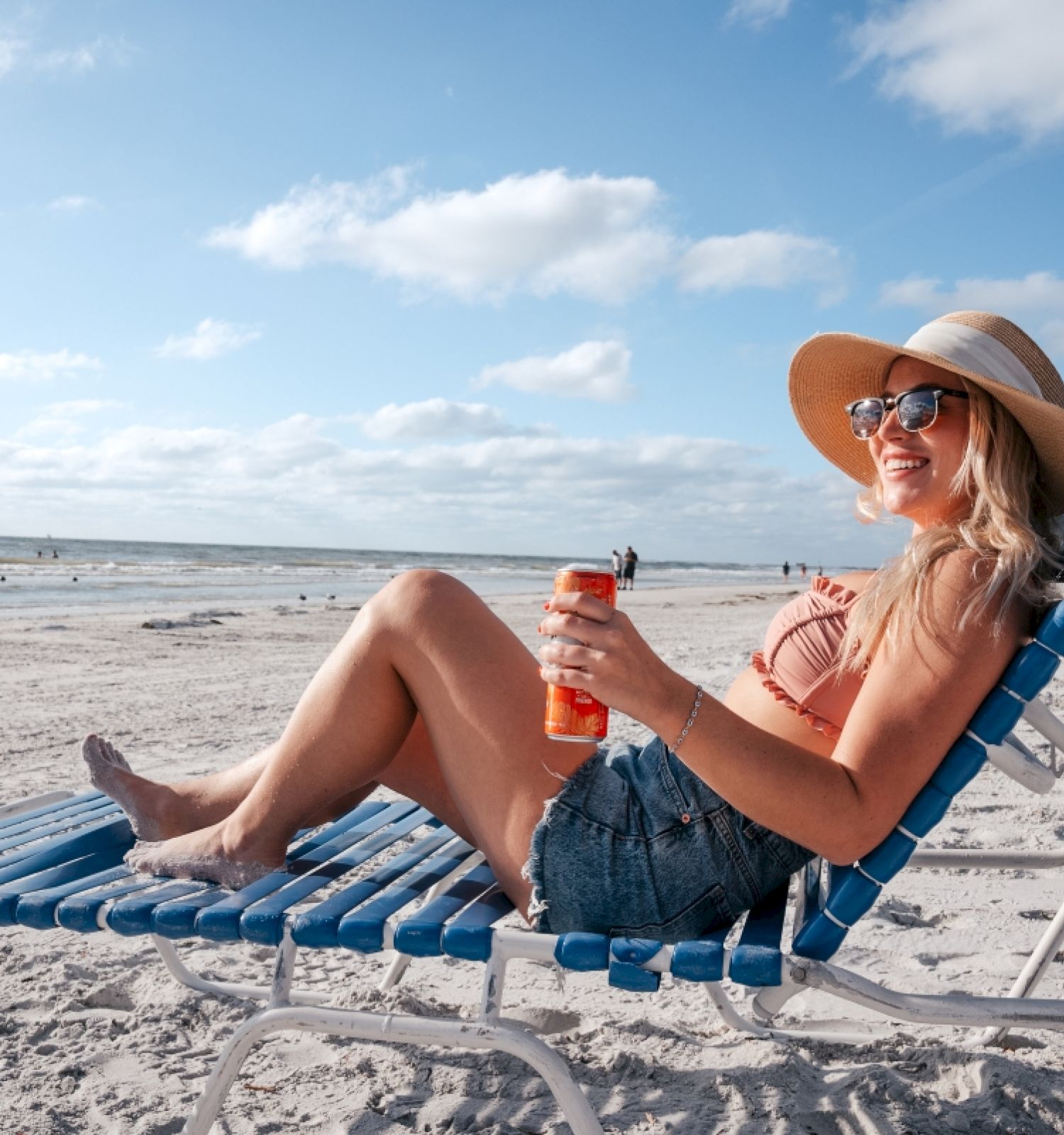 A person is relaxing on a beach chair with a drink, wearing sunglasses and a hat, on a sandy beach.