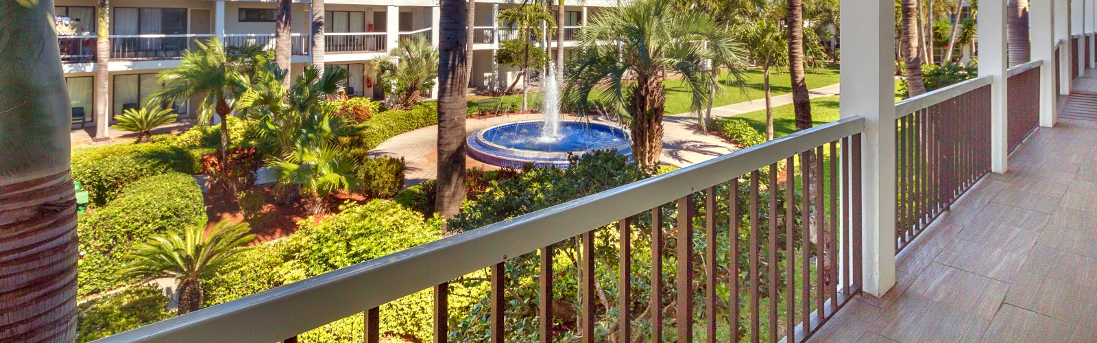 Balcony view of a tropical resort with a pool, palms, and sunny skies.