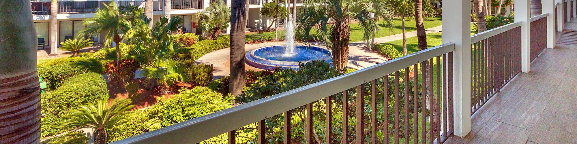 Balcony view of a tropical resort with a pool, palms, and sunny skies.