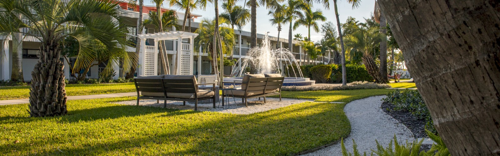 A lush garden with a fountain, palm trees, and a pathway leading to a building.