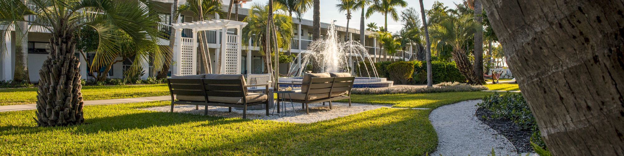 A lush garden with a fountain, palm trees, and a pathway leading to a building.