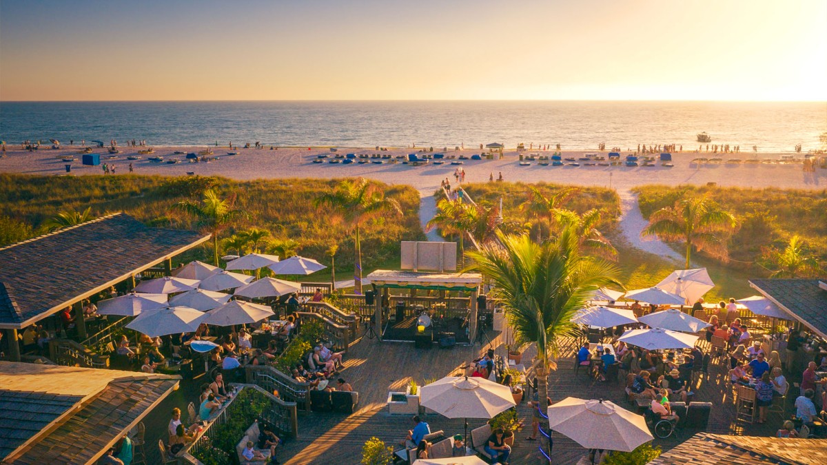 Jimmy B's on St.Pete Beach with people, umbrellas, golden sunset, and a lively atmosphere.