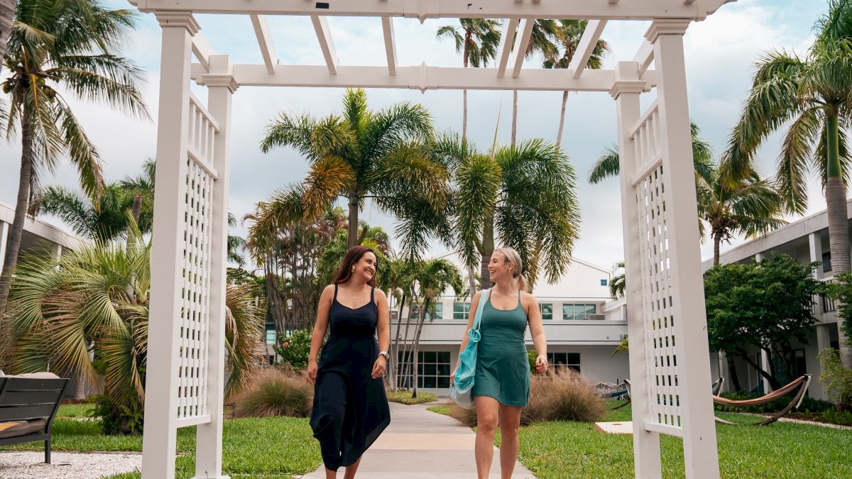 Two women walking under a pergola, surrounded by tropical landscaping.