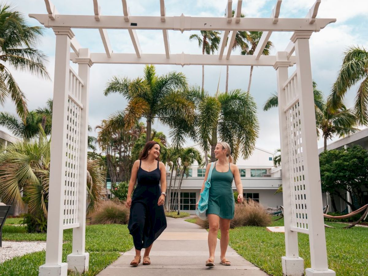 Two women walking under a pergola, surrounded by tropical landscaping.