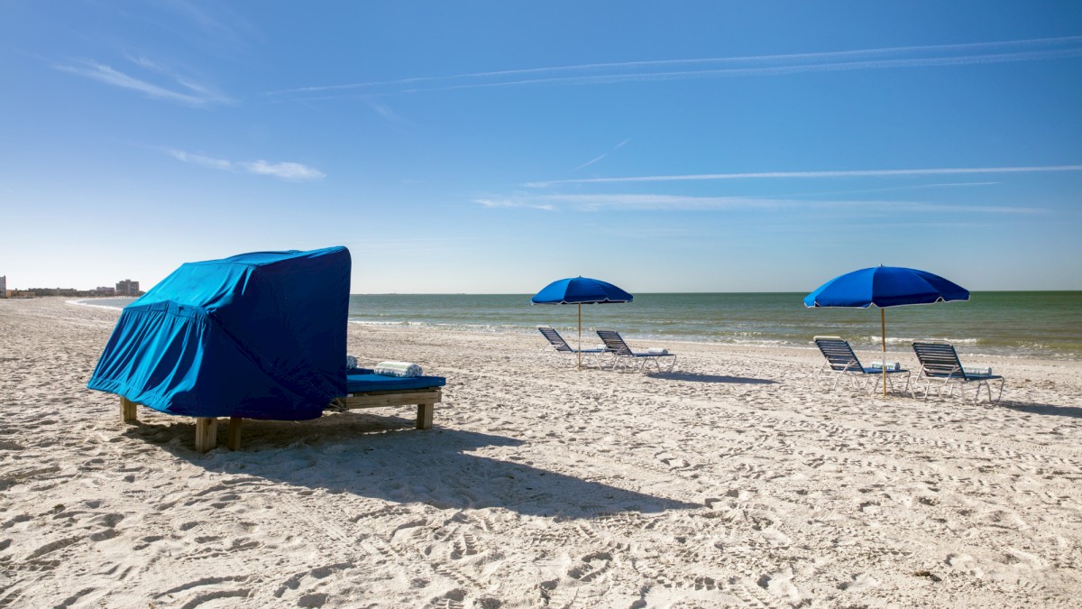 Sunny beach scene with blue umbrellas and lounge chairs on white sand.