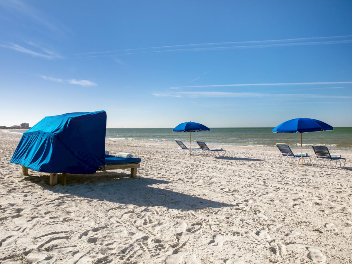 Sunny beach scene with blue umbrellas and lounge chairs on white sand.