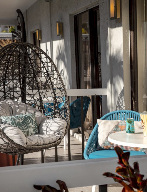 An outdoor patio with wicker chairs, a round table, and potted plants, featuring soft cushioned seating and a small standing lamp.