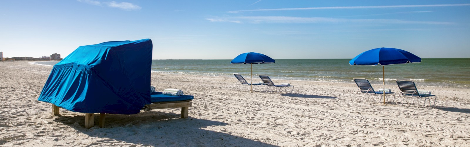 A sunny beach scene with a covered lounger and two blue umbrellas.