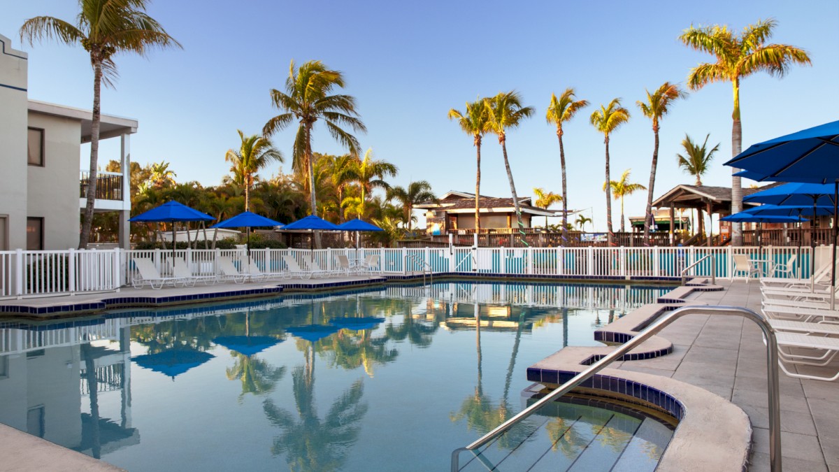 A serene pool with reflections, surrounded by palm trees and lounge chairs under blue umbrellas.