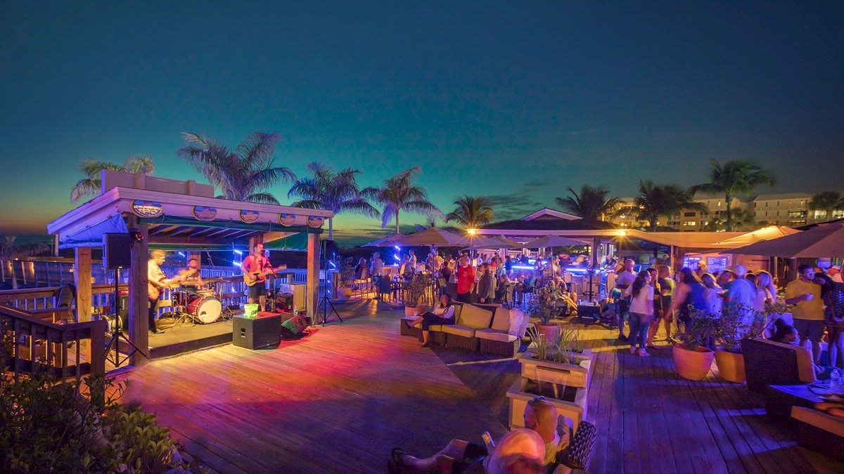 Jimmy B's beach bar at dusk with people, live music, tropical vibe, and lights.