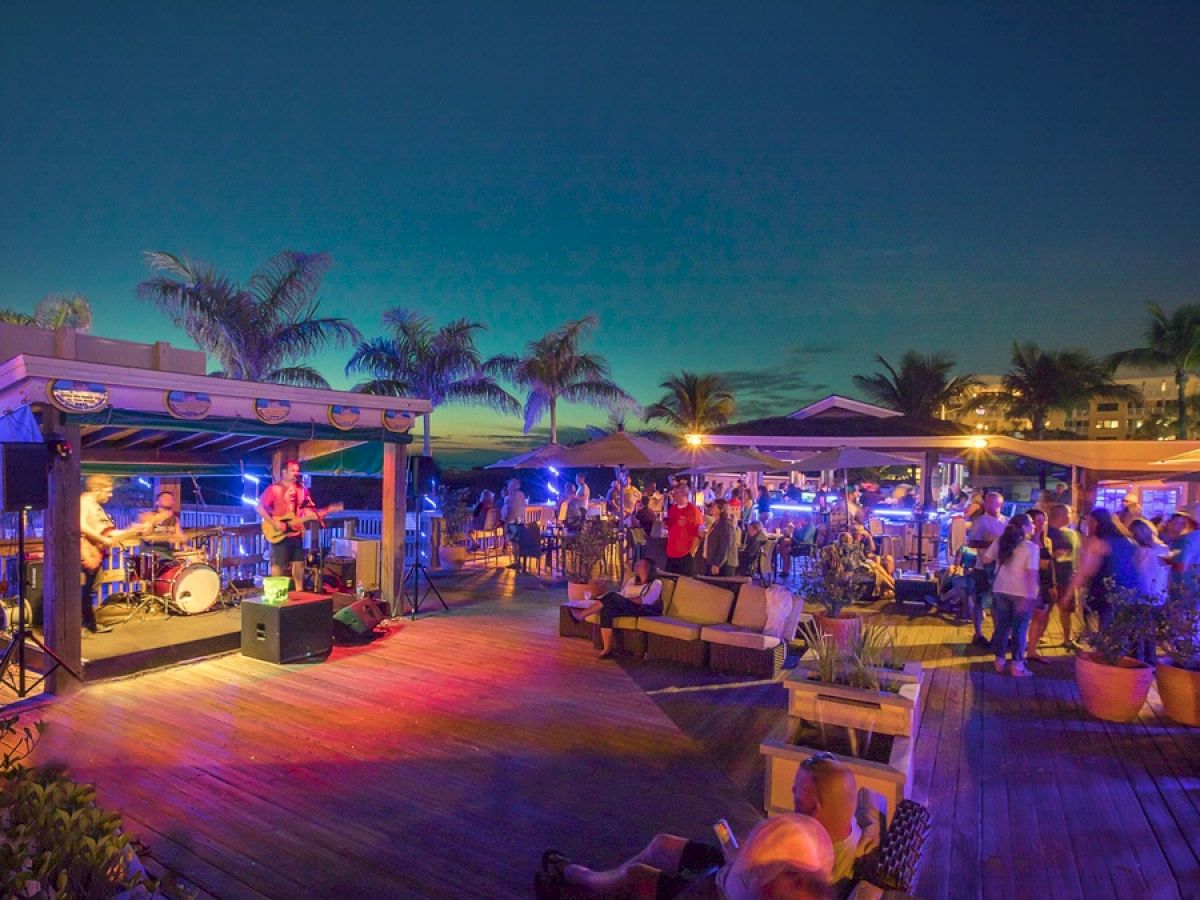 People are enjoying a vibrant outdoor evening event with live music at a beachside venue, illuminated by colorful lights and surrounded by palm trees.