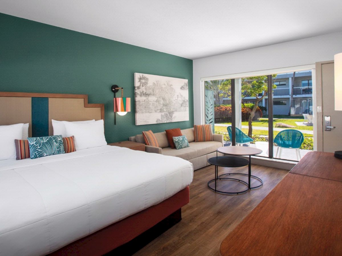 A modern hotel room features a large bed, sofa, coffee table, desk, and large window with a view of a patio and garden, creating a cozy ambiance.