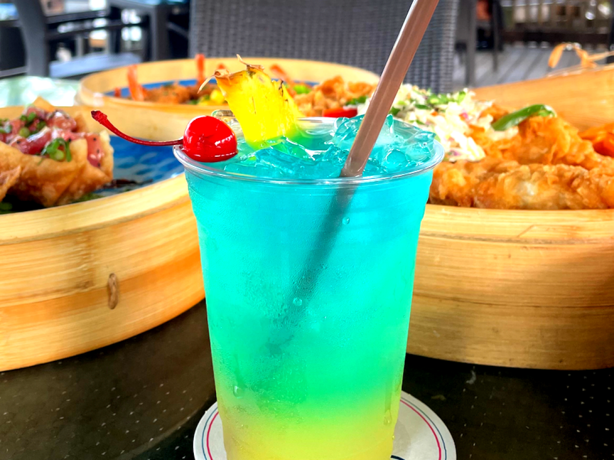 Colorful tropical drink with a cherry and pineapple slice, on a table with food in the background.