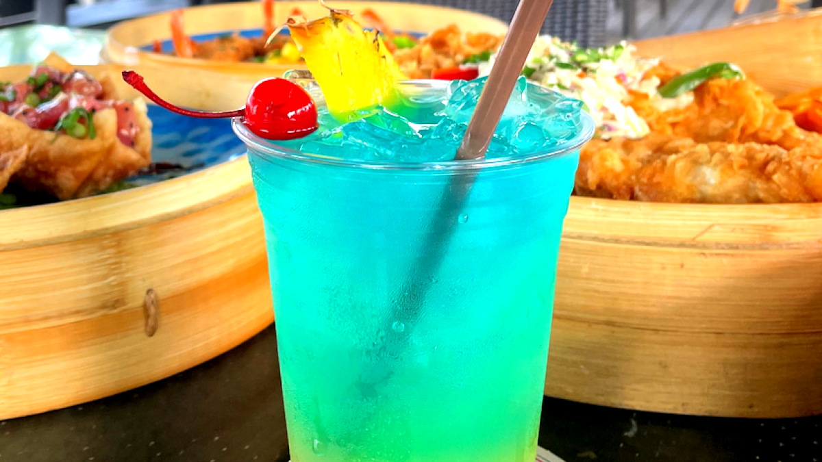 Colorful tropical drink with a cherry and pineapple slice, on a table with food in the background.