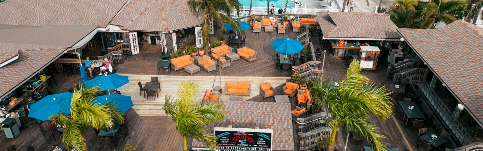 An aerial view of a tropical resort featuring outdoor seating areas with umbrellas, a swimming pool surrounded by palm trees, and several white buildings.
