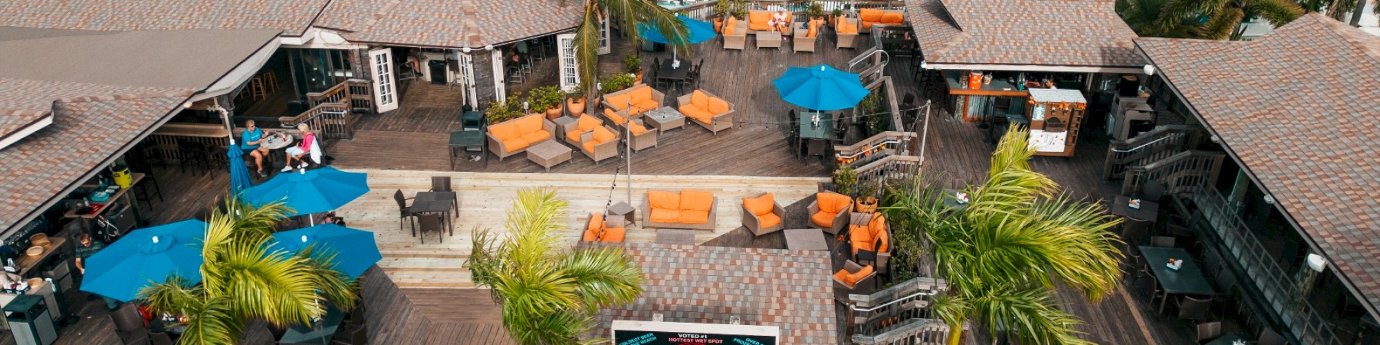 An aerial view of a tropical resort featuring outdoor seating areas with umbrellas, a swimming pool surrounded by palm trees, and several white buildings.