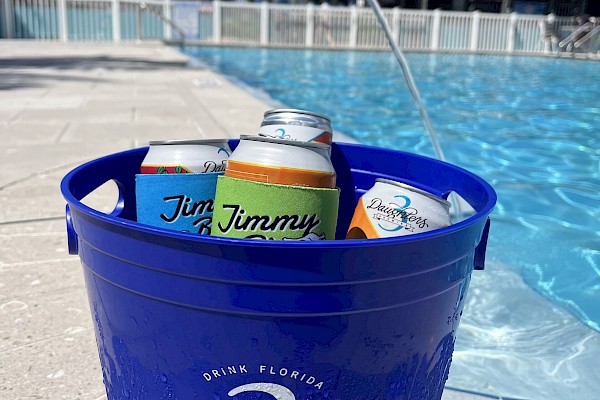 A blue bucket with cans of drinks sits on a poolside, with water splashing in the background and palm trees visible.