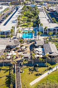 Aerial view of a resort with green spaces, two swimming pools, several buildings, and an outdoor restaurant area with tables and umbrellas.