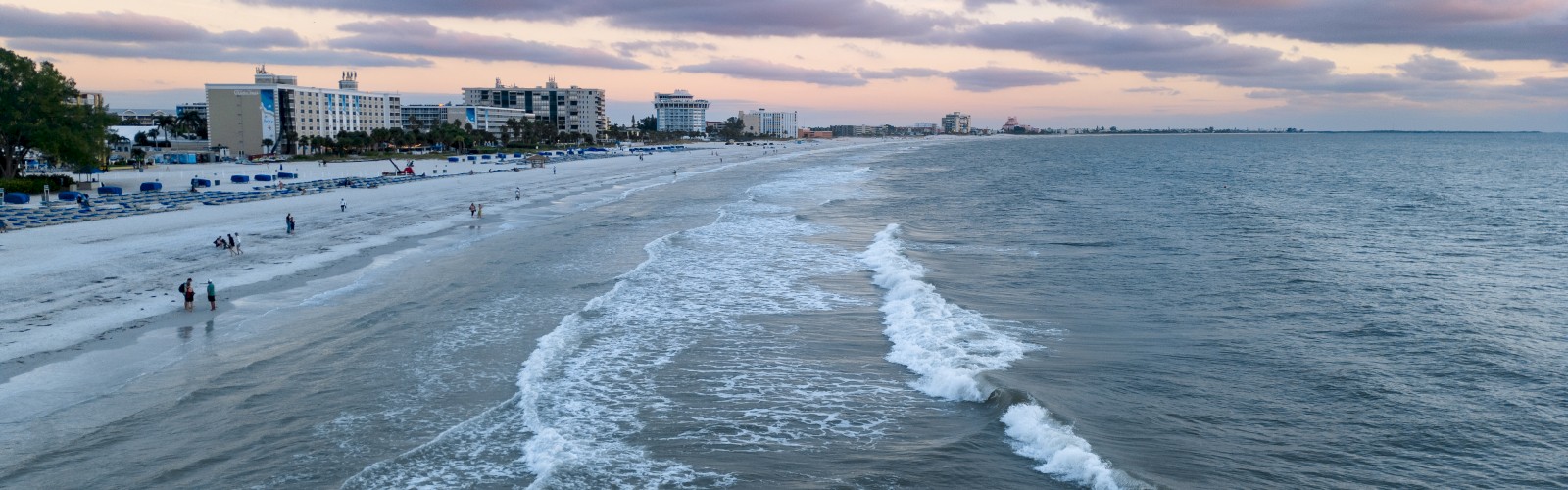 A serene beach scene at dusk with gentle waves, pink clouds, and buildings by the shore, capturing a peaceful moment in time.