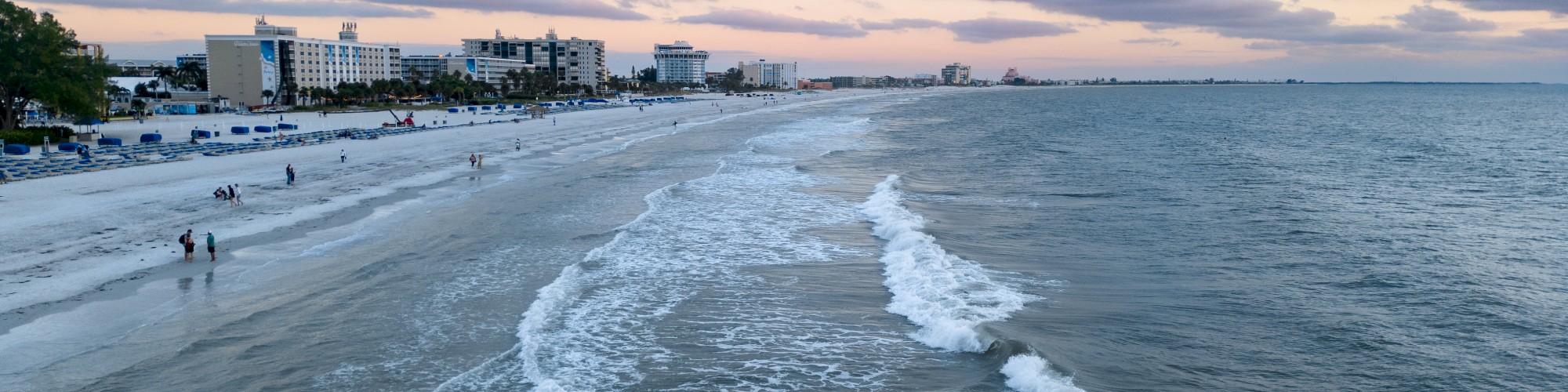 A serene beach scene at dusk with gentle waves, pink clouds, and buildings by the shore, capturing a peaceful moment in time.