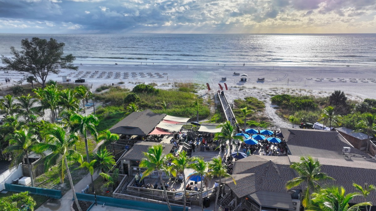 A tropical beachfront with palm trees, a resort complex by the shore, blue umbrellas, pool area, and a wooden pier leading to the ocean, at sunset.