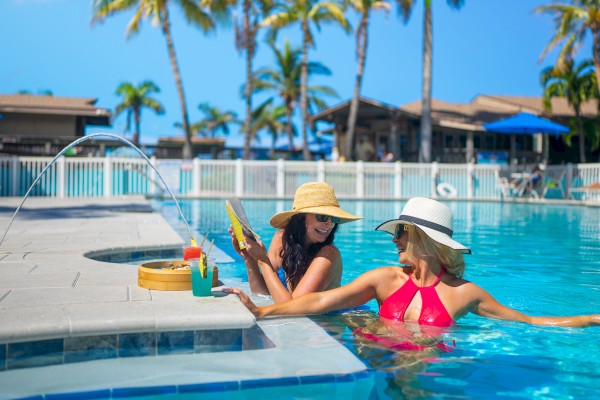 Two people relaxing in a pool with drinks nearby, surrounded by palm trees and lounge chairs, enjoying a sunny day.