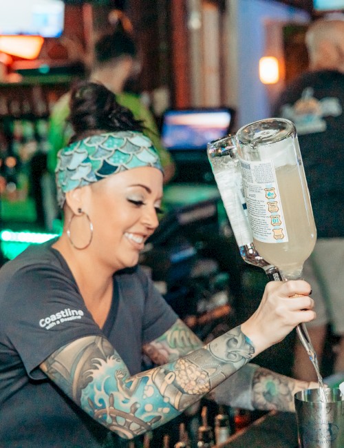 A bartender with tattoos pours a drink behind a bar with bottles and other patrons in the background.