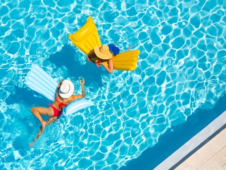 Two people relax on inflatable mats in a pool, with a palm tree shadow on the surrounding deck.