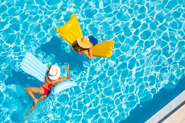 Two people relax on inflatable mats in a pool, with a palm tree shadow on the surrounding deck.