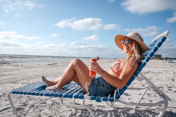 A person is relaxing on a beach chair, wearing a sunhat and sunglasses, holding a drink. The beach and ocean are in the background.