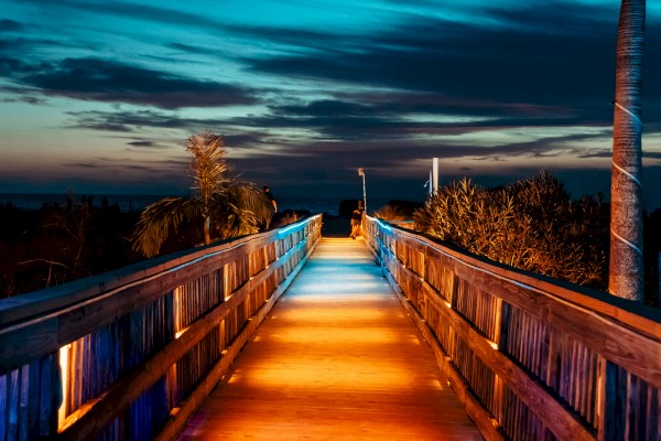 A wooden pathway illuminated by warm lights leads towards a scenic view, surrounded by palm trees under a vibrant evening sky.