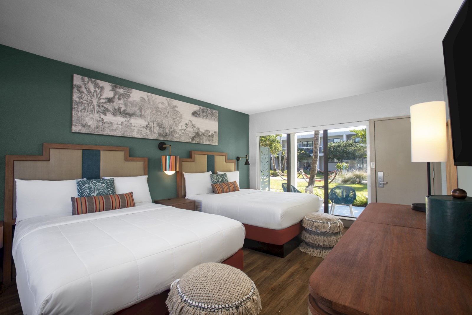 The image shows a hotel room with two beds, a TV, wooden furniture, a decorative wall art, and a view of a garden through glass doors.