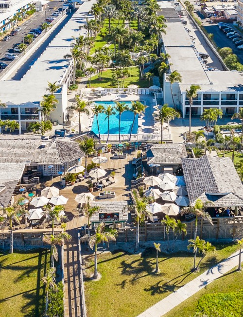 Aerial view of a resort area with multiple buildings, a pool, and outdoor seating surrounded by palm trees and grassy areas.