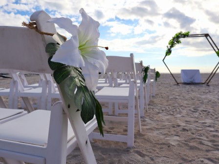 A beach wedding setup with white chairs, floral decorations, and a hexagonal arch adorned with greenery, set against a cloudy sky.