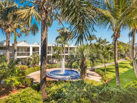A lush garden scene with palm trees surrounds a building and a fountain at its center, under a clear blue sky.