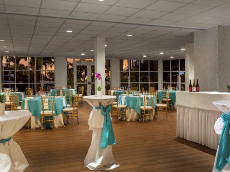 The image shows a decorated event space with tables, turquoise accents, tall tables with flowers, and a bar area with bottles.