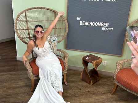 A woman in a white lace wedding dress sits in a wicker chair, posing with one arm raised near a round chair, in a bright room next to a sign.