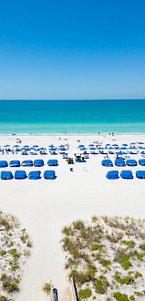 The image shows a beach with rows of blue umbrellas and chairs on white sand, with the ocean and a few people in the background.