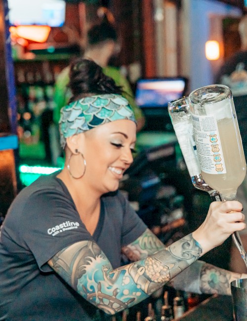 A bartender with tattoos and a patterned headband is pouring a drink. Various bottles and bar tools are visible in the background.