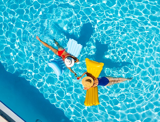 A person in a yellow life vest floats on a bright blue pool, wearing a hat, with a yellow paddleboard or float beside them.