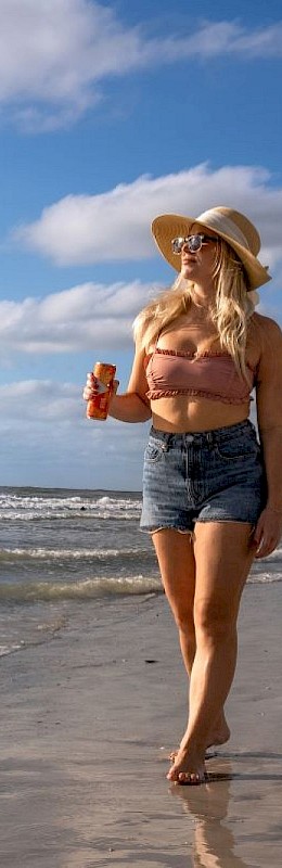 A woman in a swimsuit walks on a sunny beach, waves near the shore, blue sky above, onlookers in distance, bright and cheerful.