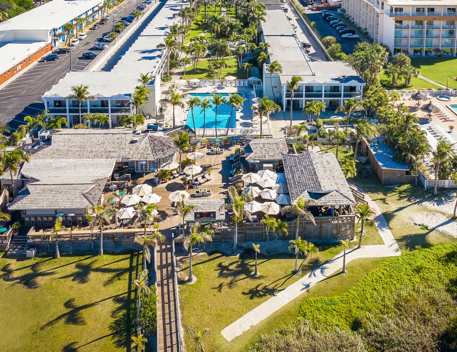 The image shows a resort complex with a pool, surrounded by palm trees, pathways, and various buildings, in a sunny setting.