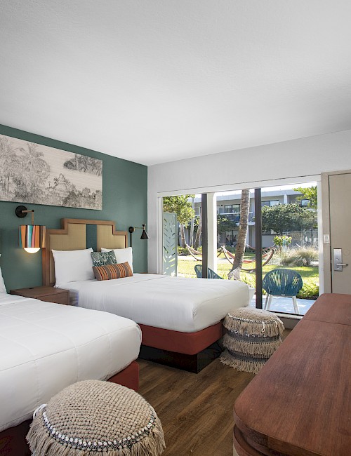 Two double beds in a modern hotel room with a teal accent wall, wall art, a wooden desk, ottomans, and a sliding glass door to a patio.