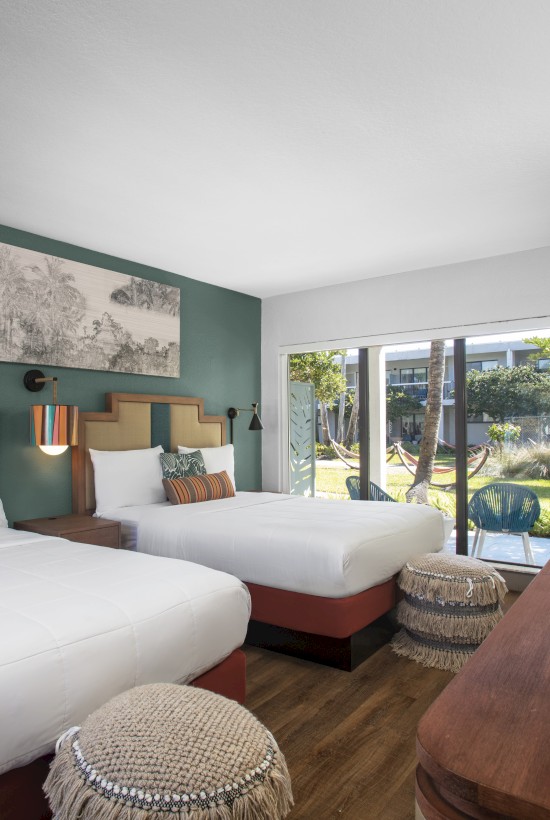 A hotel room with two beds, decorative pillows, a large window, wooden furniture, and a landscape artwork on the wall.