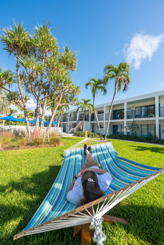 A person relaxing on a striped hammock in a sunny hotel courtyard with palm trees and a white two-story building in the background.