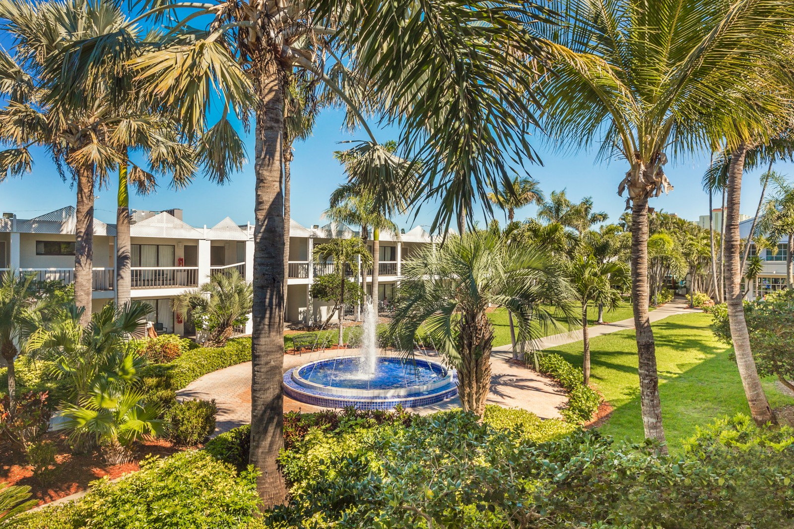 A tropical resort courtyard with palm trees, a circular blue fountain in the center, green lawns, and white cottages in the background.