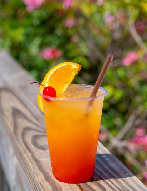 A tropical sunset drink in a plastic cup with orange to red gradient, garnished with an orange slice and cherry on a wooden railing. Ends with a period.