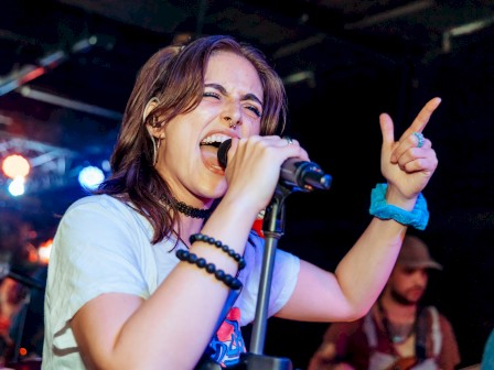 A female singer performs on stage, holding a microphone with her left hand raised in a rock gesture, wearing bracelets and a white tee.