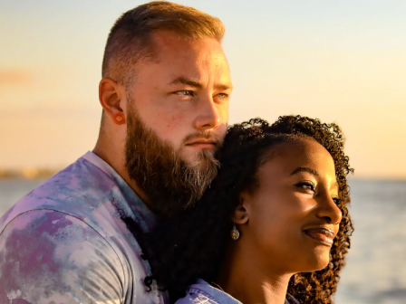 A couple stands close by the ocean at sunset, cuddling with calm smiles, the man with a beard and the woman with curly hair.